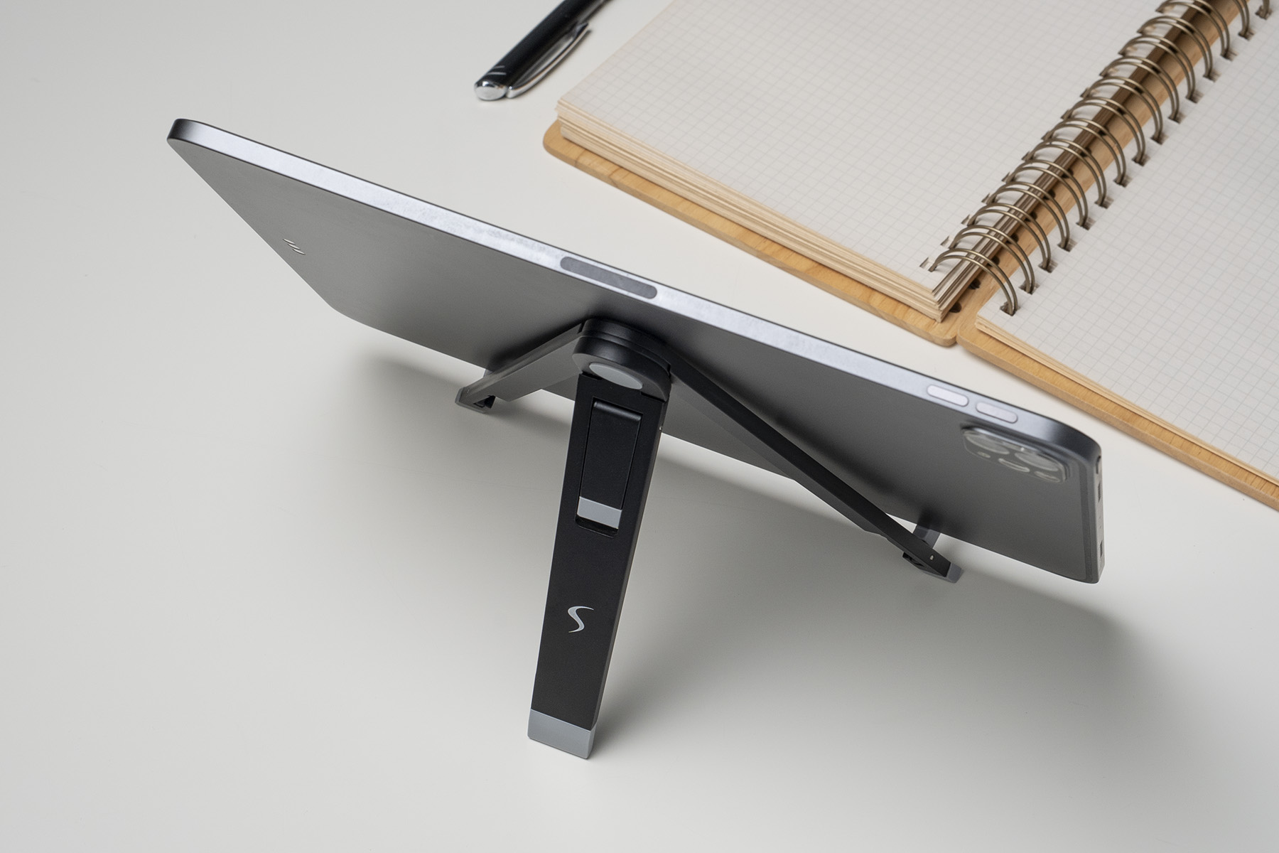 Ein Tablet wird von einem Ständer auf einer weißen Oberfläche gehalten, mit einem Notizbuch und Stift im Hintergrund.