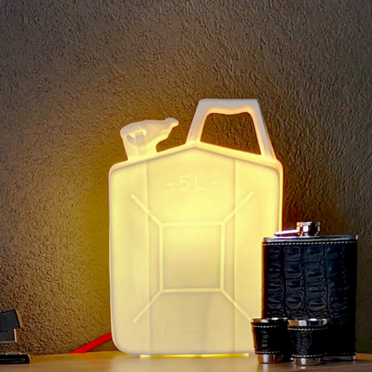 Een gloeiende jerrycan lamp staat op een tafel. Een fles en borrelglaasjes staan ernaast. Donkere muur op de achtergrond.
