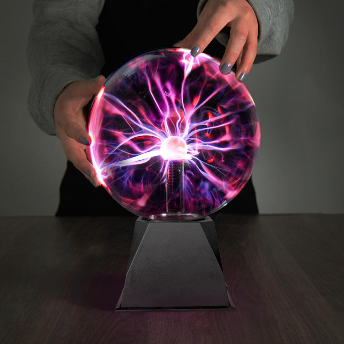 Le mani di una persona tengono una sfera al plasma che emette archi elettrici viola e rosa. La sfera poggia su un supporto nero su una superficie di legno.