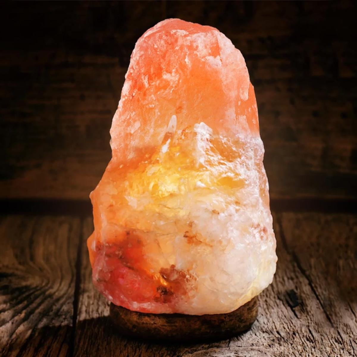Een gloeiende Himalaya zoutlamp met een oranje tint staat op een houten voet tegen een donkere houten achtergrond.