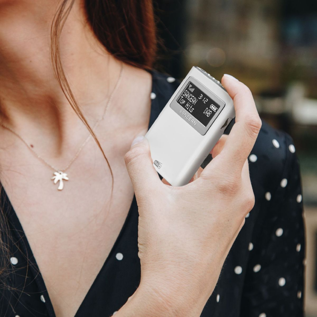 Kobieta trzyma białe radio z godziną i 'SANGEAN Top Hits'. Ma na sobie czarną koszulę w kropki i naszyjnik z palmą.