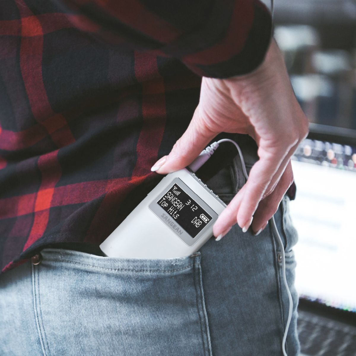 Osoba wkłada białe radio do kieszeni dżinsów. Ekran radia pokazuje 'SANGEAN Top Hits DAB'. Ma na sobie koszulę w kratę.