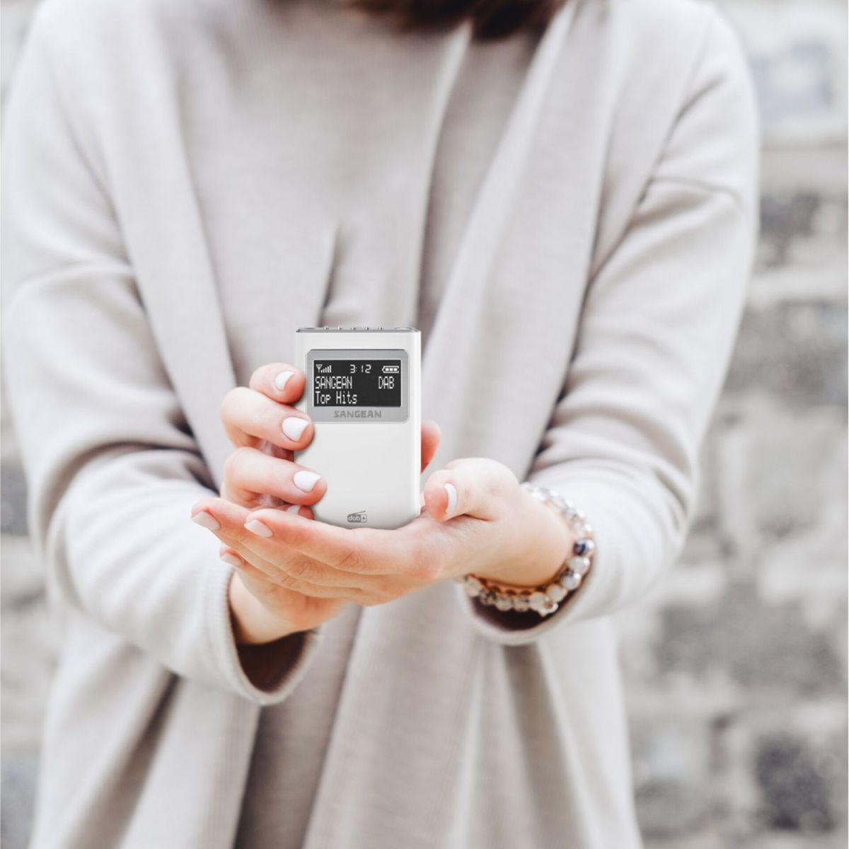 Osoba trzyma białe radio SANGEAN. Ekran wyświetla 'SANGEAN Top Hits'. Osoba ma na sobie beżową kurtkę i bransoletkę.