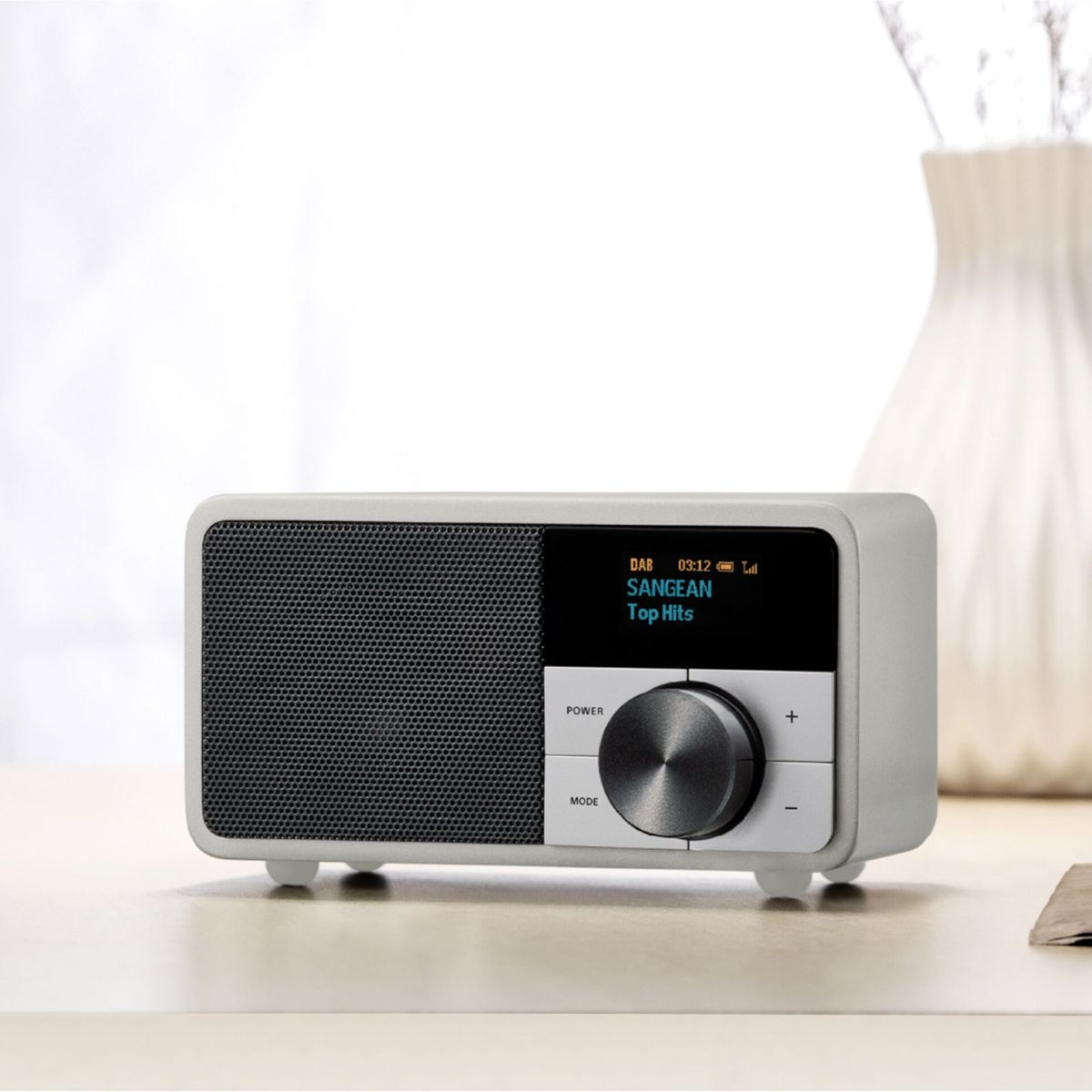 Une radio grise avec un écran noir affichant 'SANGEAN Top Hits'. Elle a un bouton et des boutons, posée sur une table claire.