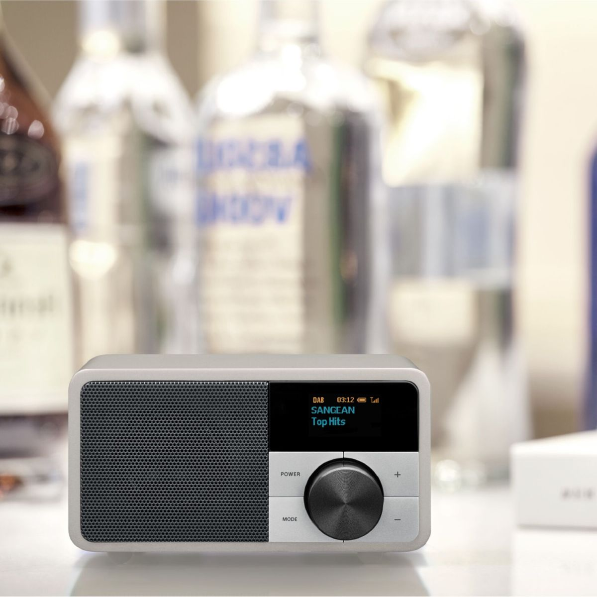 Une radio grise avec une grille de haut-parleur noire et un écran. Bouteilles d'alcool floues en arrière-plan.