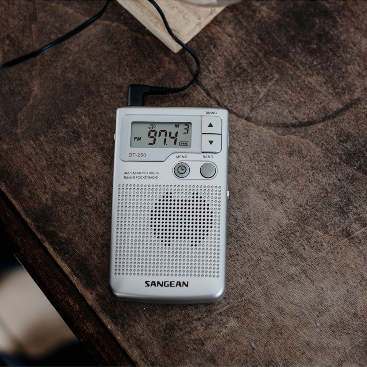 Srebrne radio kieszonkowe SANGEAN wyświetla 97,4 FM na drewnianej powierzchni. Czarny kabel jest podłączony. Radio ma przyciski i ekran.