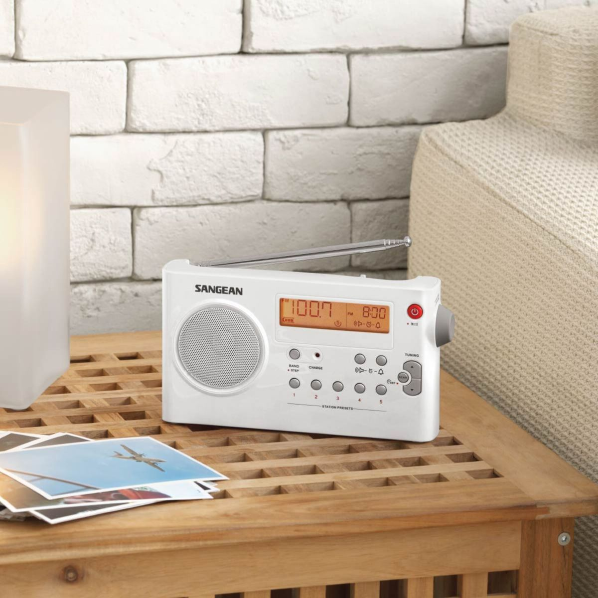 Ein weißes Sangean-Radio steht auf einem Holztisch mit Fotos. Im Hintergrund sind eine Lampe und ein beiges Sofa. Eine Backsteinmauer ist dahinter.