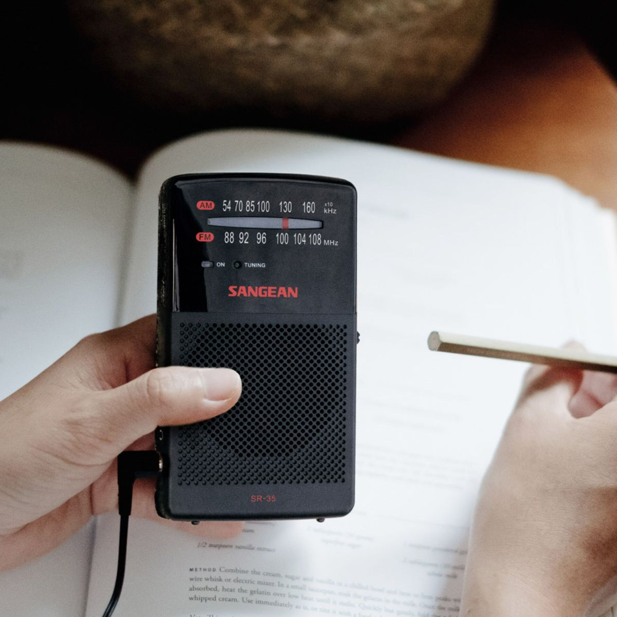 Una persona sostiene una radio Sangean negra, un lápiz descansa sobre un libro abierto y el fondo está borroso.