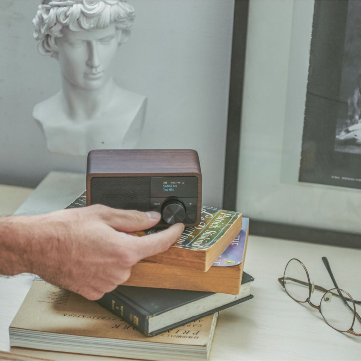A hand adjusts a wooden radio on books. A bust and framed art are in the background. Glasses lay on the table.