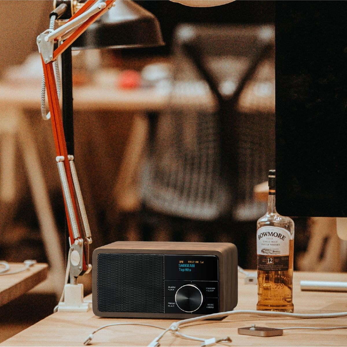 A wooden radio sits on a desk next to a lamp and a bottle of whisky. The radio's screen displays 'SANGEAN Top Hits'.