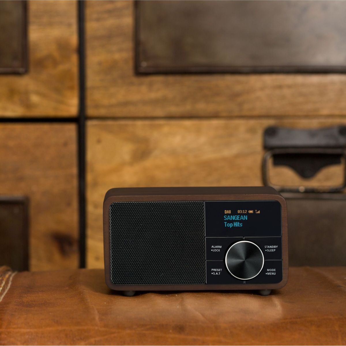 A brown wooden radio with a black speaker. The screen displays 'SANGEAN Top Hits'. Buttons include alarm, preset, standby, and mode.