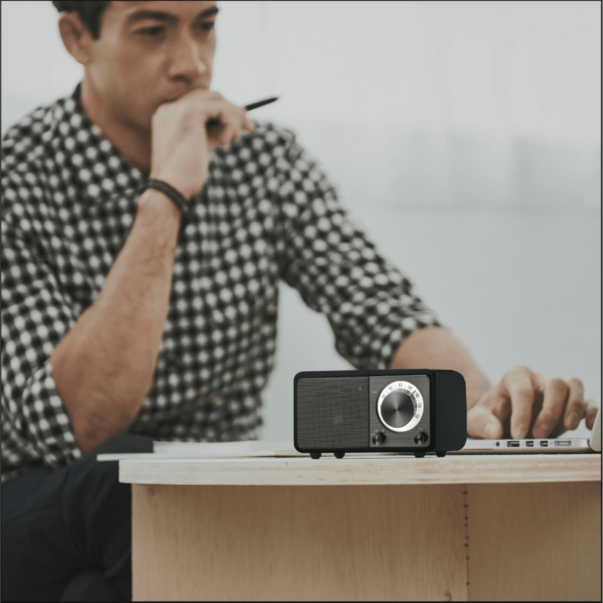 Mężczyzna w koszuli w kratę siedzi przy biurku z radiem i laptopem. Trzyma długopis, wyglądając na zamyślonego.