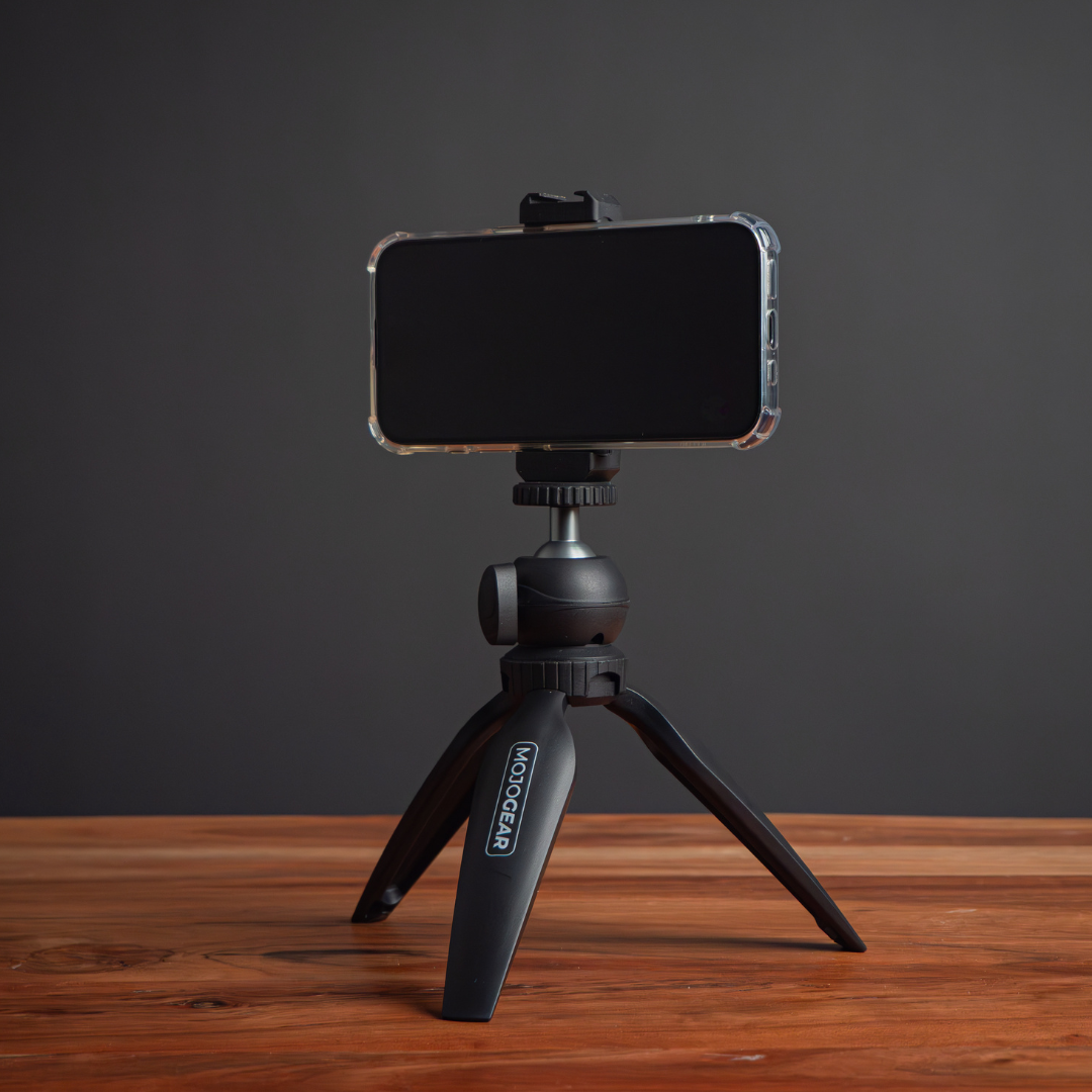 A smartphone is mounted on a black tripod. The tripod sits on a wooden surface against a dark gray background.