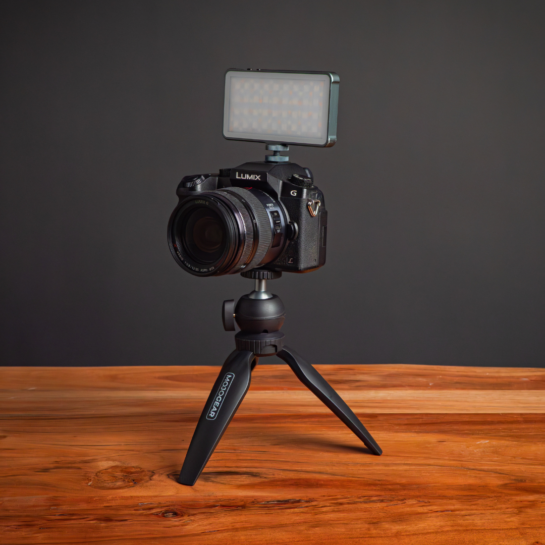A camera with a light is mounted on a tripod. The setup is on a wooden surface with a dark gray background.