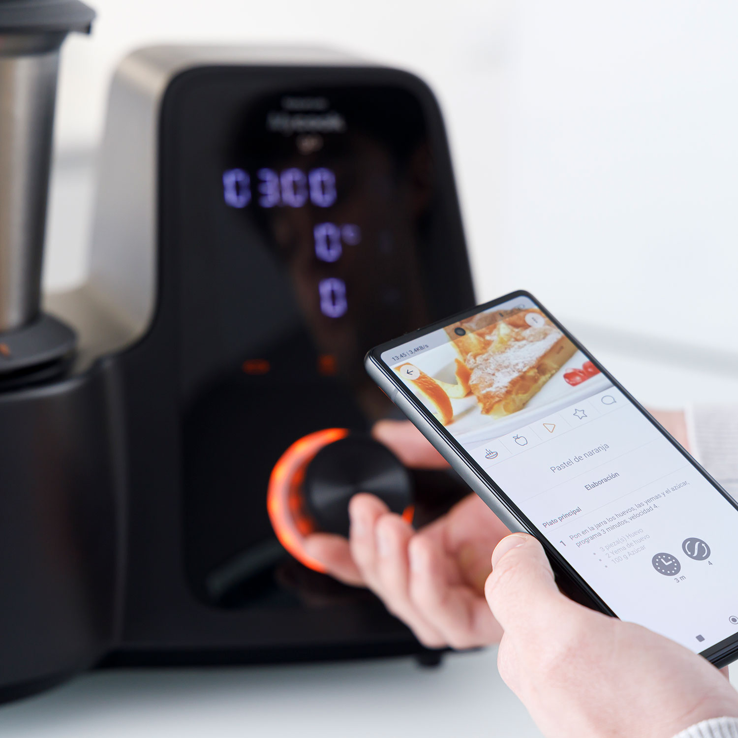 Una persona sostiene un teléfono que muestra una receta de pastel de naranja. Un procesador de alimentos está en el fondo, con un dial iluminado en naranja.