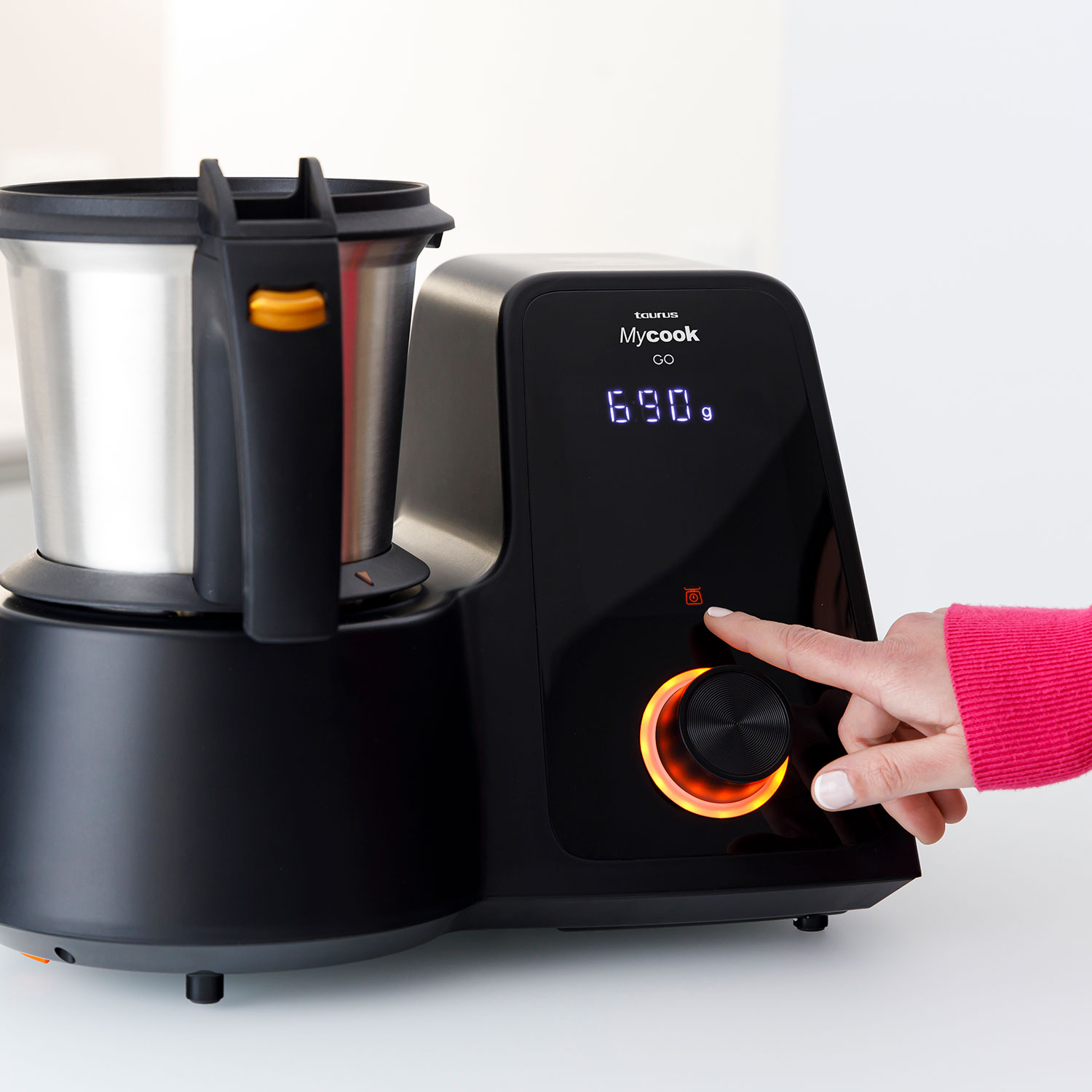 Un procesador de alimentos negro Taurus Mycook Go. Una mano presiona el botón de inicio. La pantalla muestra 690g. El fondo es blanco.