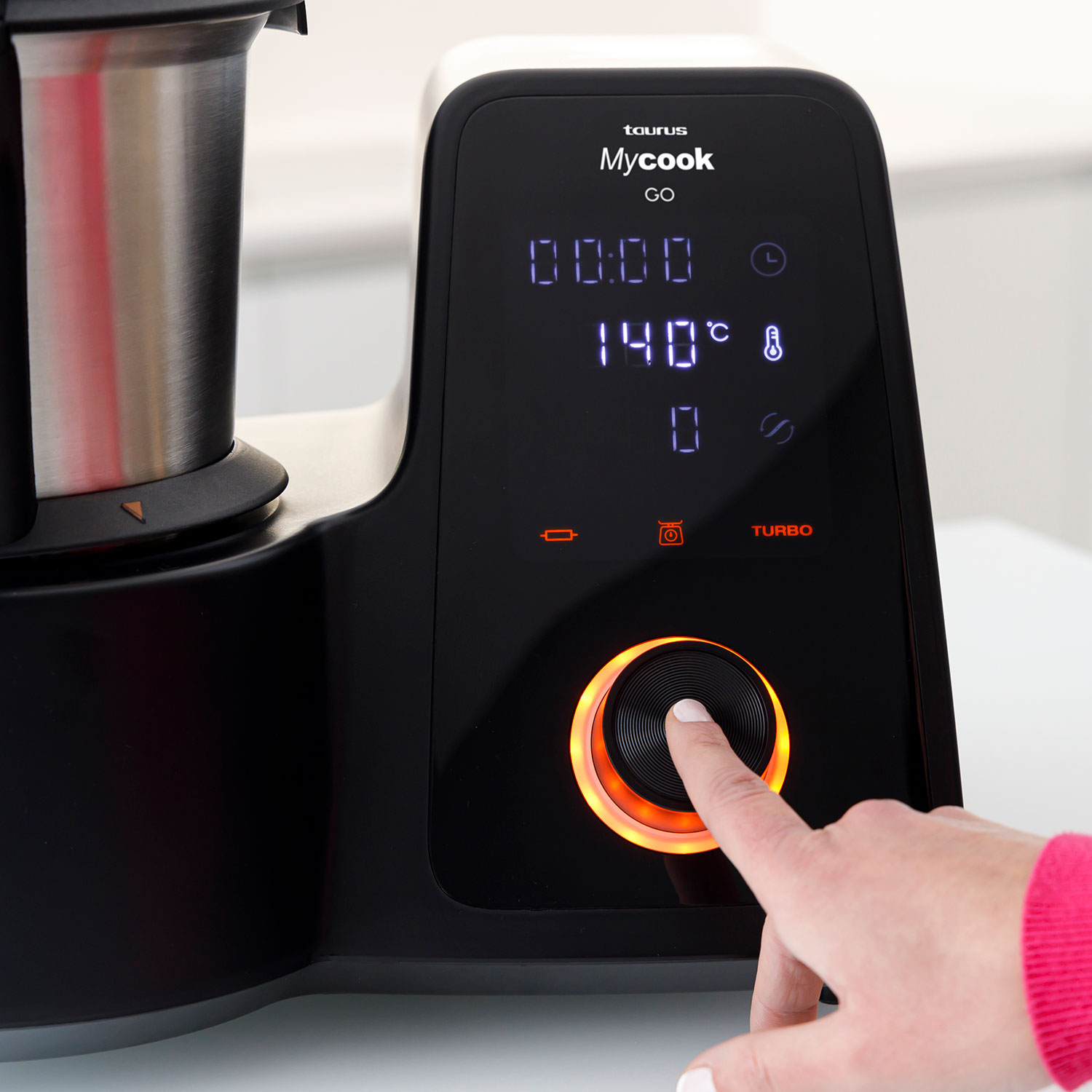 Una mano presiona un botón en un Taurus Mycook GO negro. La pantalla muestra 140°C. El botón brilla en naranja.