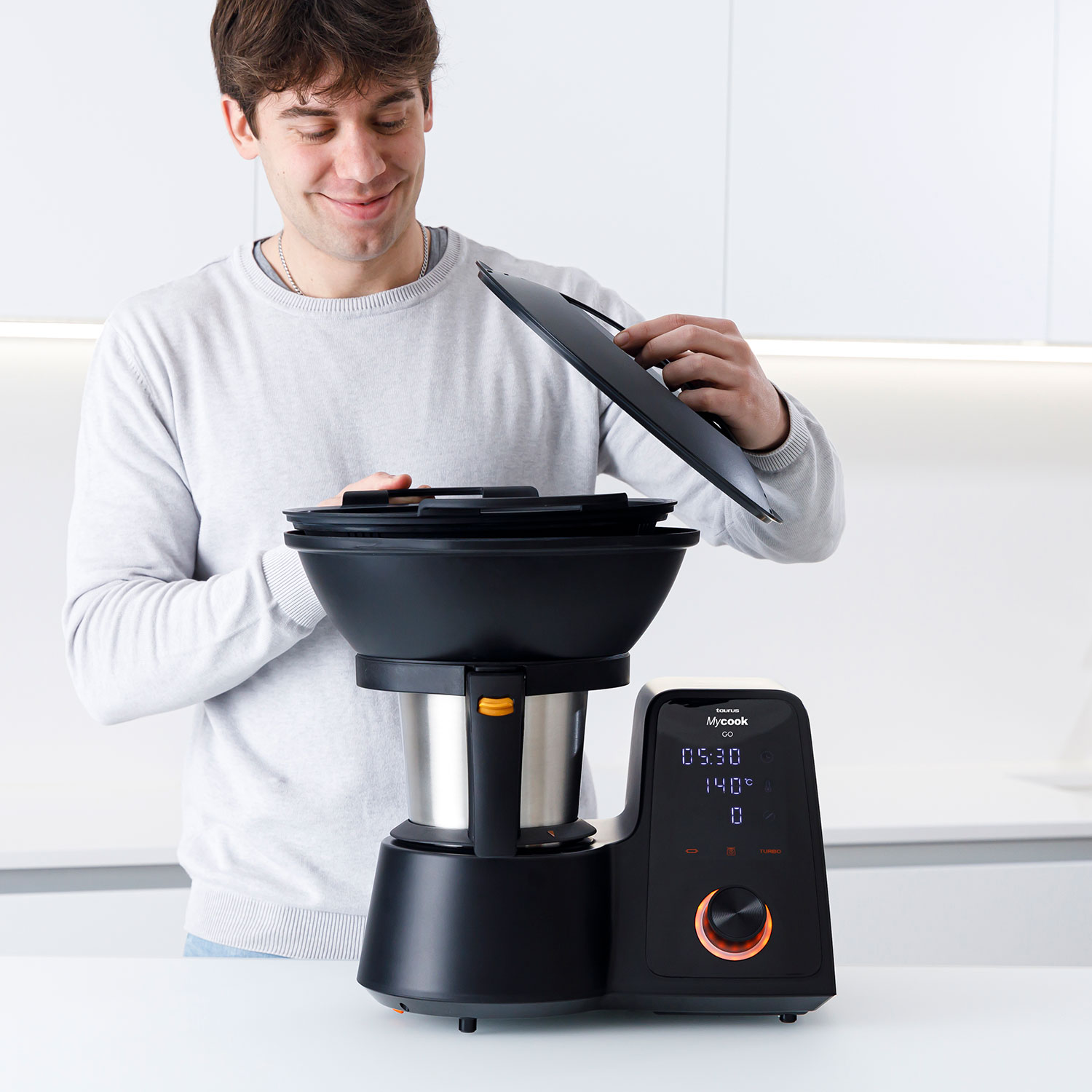 Un hombre abre un procesador de alimentos negro. Tiene una pantalla digital que muestra la hora y la temperatura. Una cocina blanca es el fondo.