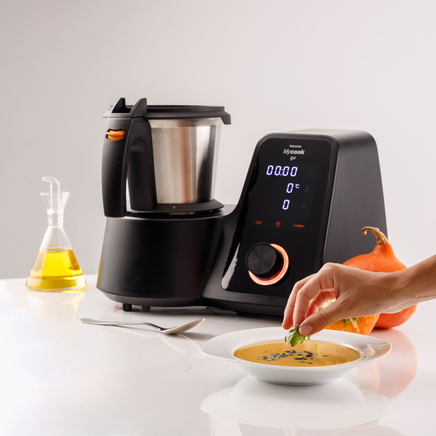 Un procesador de alimentos junto a un cuenco de sopa. Una mano espolvorea ingredientes. En la mesa hay una calabaza y una botella de aceite.