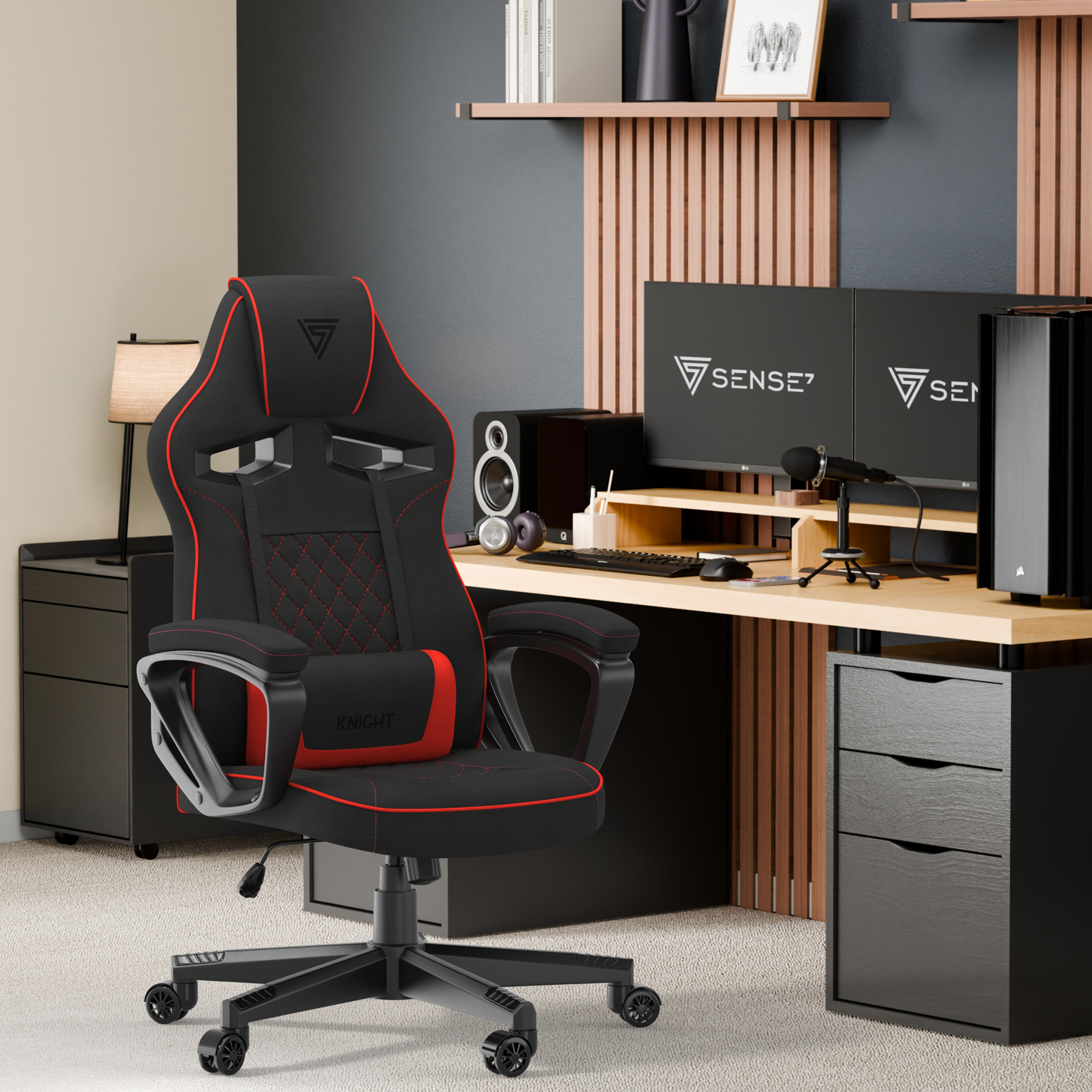 Configuration de jeu avec chaise noire et rouge, deux moniteurs, bureau, haut-parleurs et accessoires. Une lampe et des étagères sont visibles.
