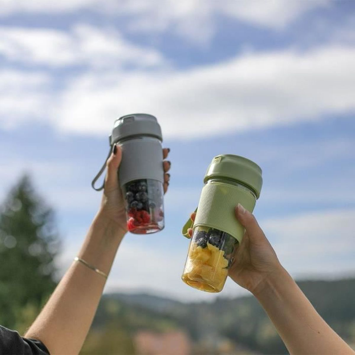 Zwei Hände halten Mixer mit Früchten vor blauem Himmel. Einer hat Beeren, der andere Ananas.