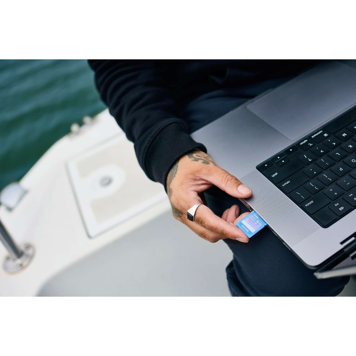 Eine Person steckt eine blaue SD-Karte in einen Laptop. Die Person trägt einen Ring und hat Tätowierungen auf der Hand. Der Hintergrund ist unscharf.