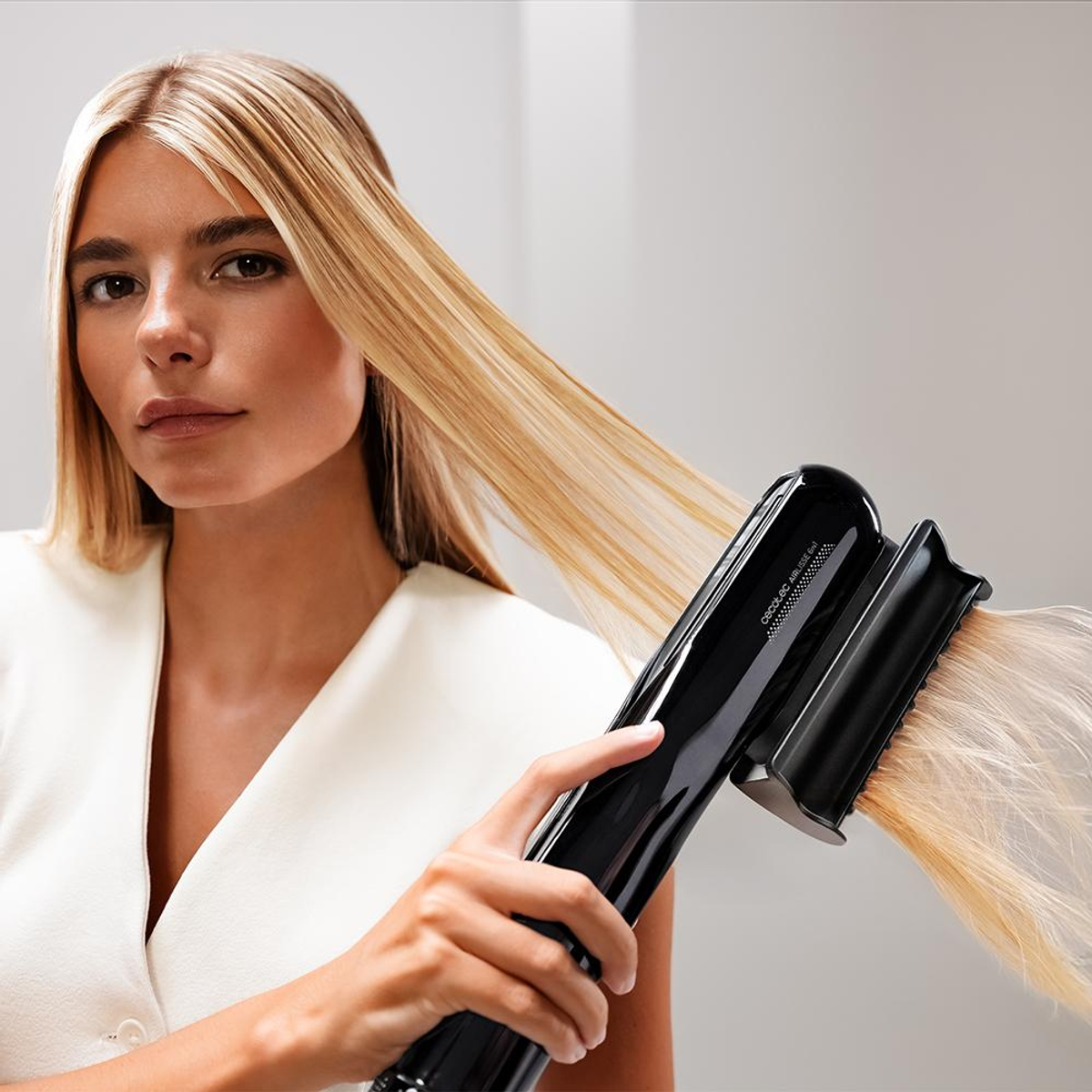 Kobieta z blond włosami używa czarnej prostownicy. Ma na sobie biały top. Tło to gładka biała ściana.