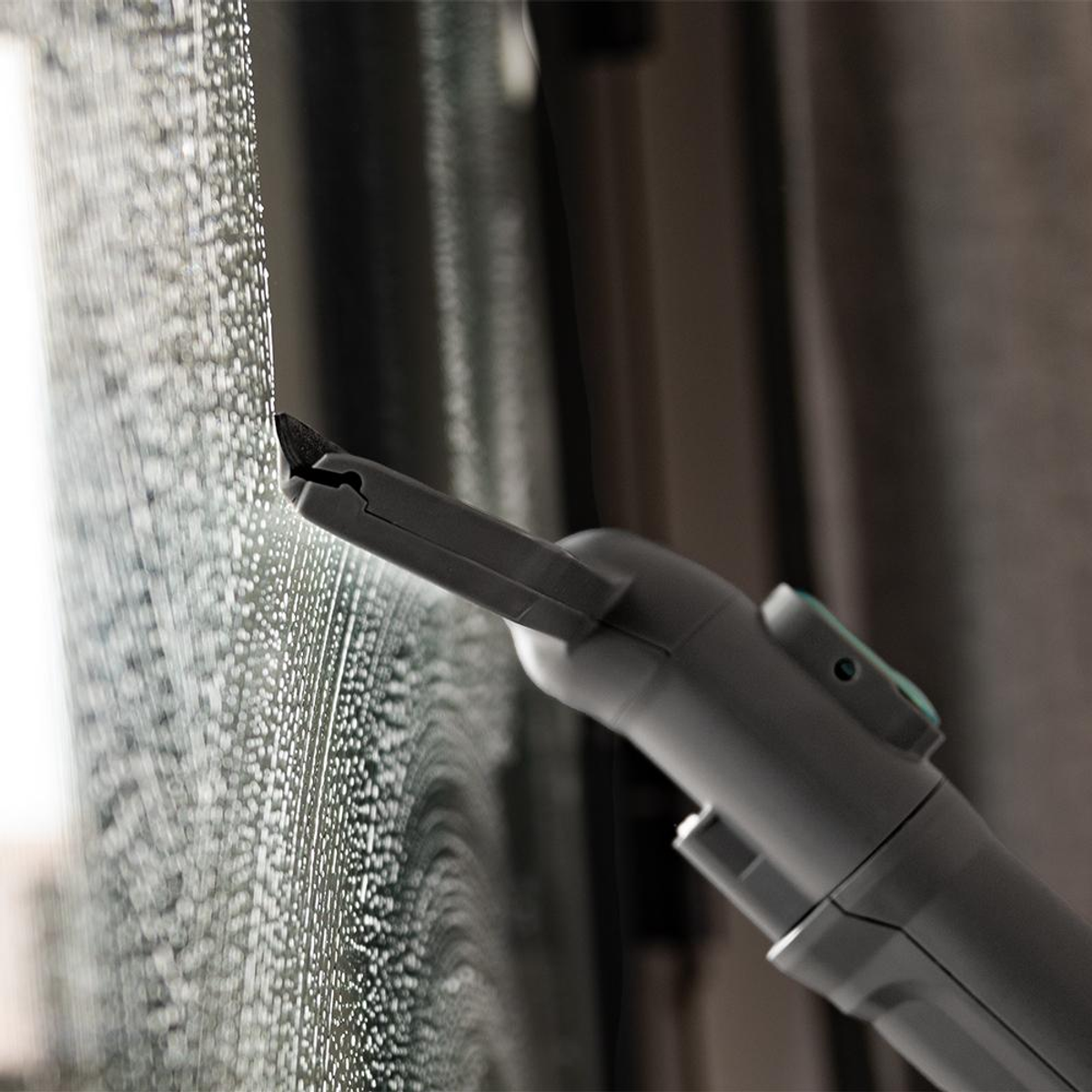 Una herramienta gris para limpiar ventanas está limpiando una ventana jabonosa. La ventana tiene muchas burbujas pequeñas. El fondo está borroso con cortinas.