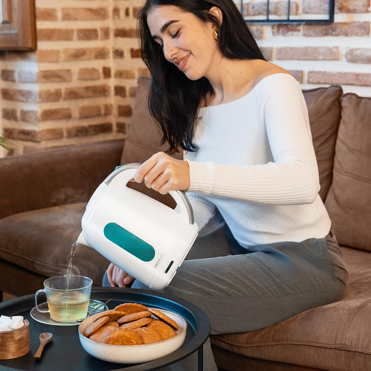 Una donna versa l'acqua da un bollitore bianco in una tazza di tè di vetro. Biscotti e una zuccheriera sono su un tavolo.