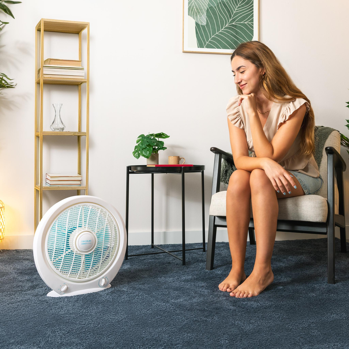 Een vrouw zit lachend op een stoel. Een ventilator staat voor haar. Een tafeltje met een plant links van haar. Een boekenrek staat achter haar.