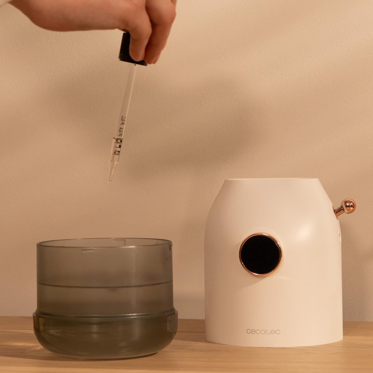 Eine Hand hält eine Pipette über einem grauen Behälter und einem weißen Luftbefeuchter. Die Pipette tropft Flüssigkeit.