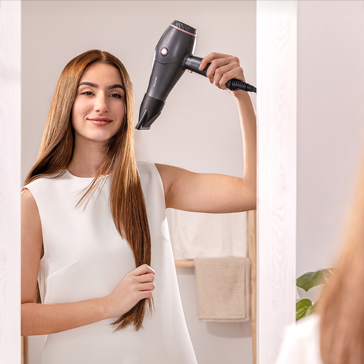 Una mujer sostiene un secador de pelo. Viste un vestido blanco, tiene pelo castaño largo y sonríe. El secador es negro y rosa.