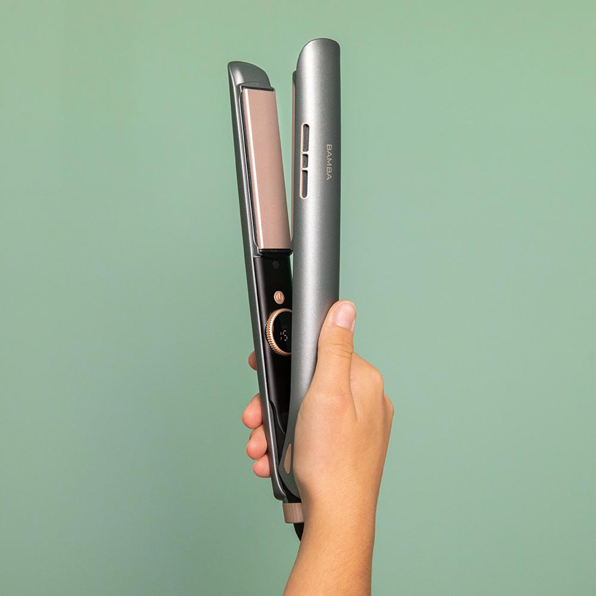 A hand holds a silver and black hair straightener. The background is a solid green color.