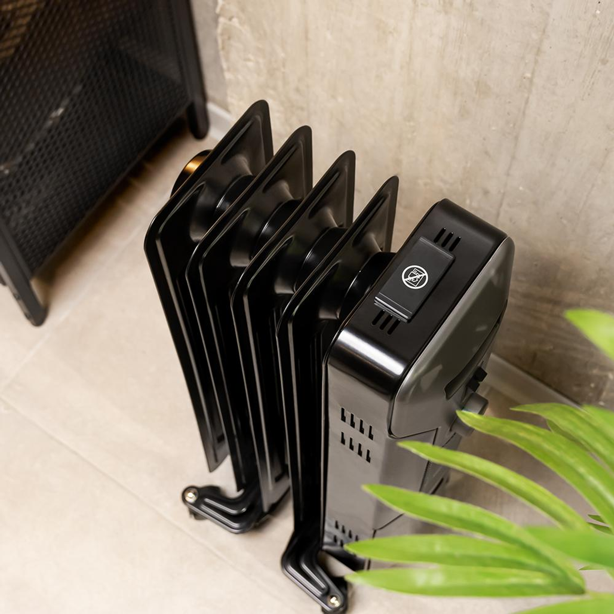 Czarny grzejnik olejowy stoi na jasnej podłodze. Zielone liście roślin z przodu, z czarnym metalowym przedmiotem w tle.