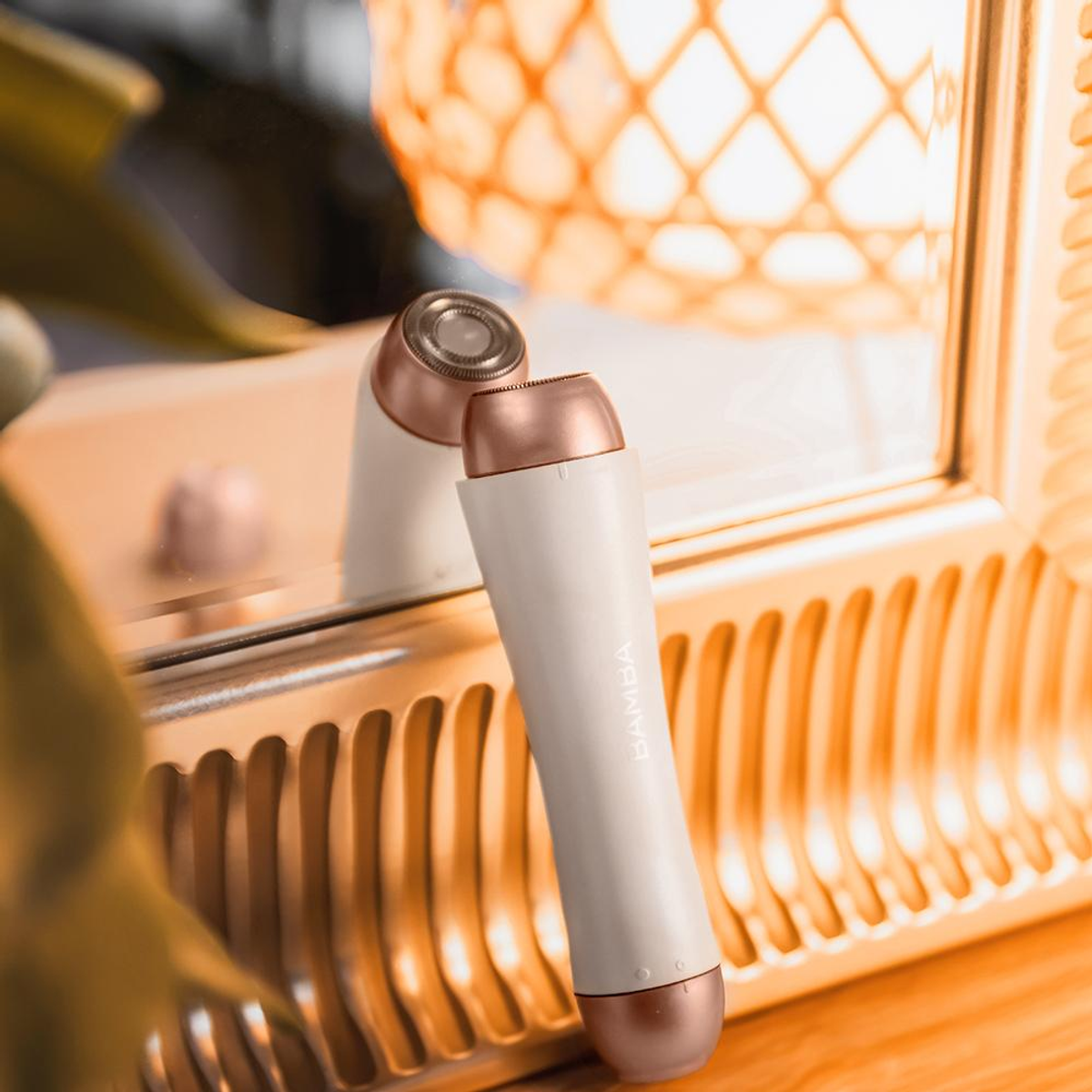 Una afeitadora eléctrica blanca y oro rosa está sobre un espejo con marco dorado. El fondo muestra una lámpara decorativa.