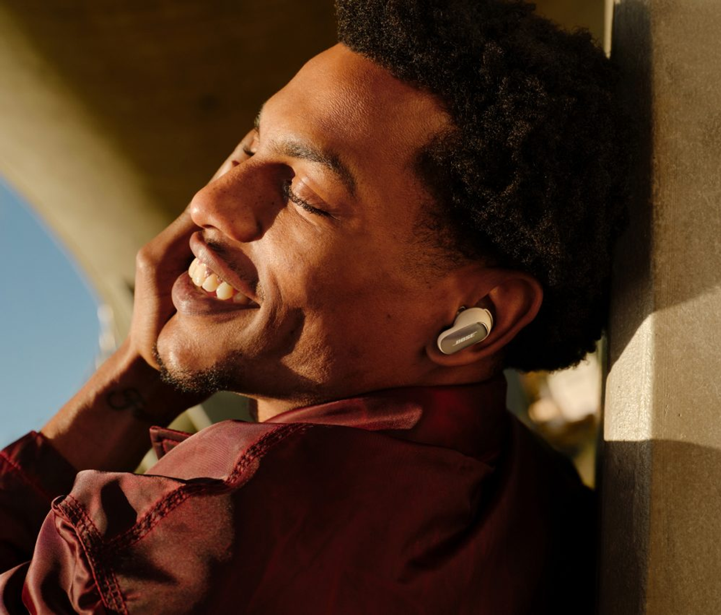 Hombre negro sonriente con auriculares, apoyado en una pared. Lleva una camisa granate y tiene el pelo rizado. Fondo soleado.