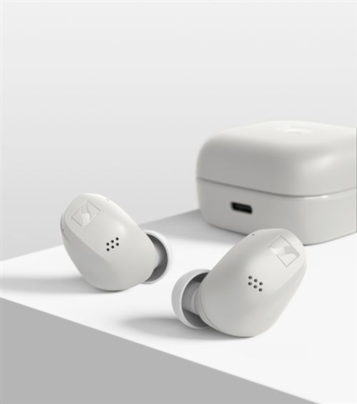Dos auriculares inalámbricos blancos y un estuche de carga sobre una superficie blanca. Los auriculares tienen el logo de la marca y pequeños agujeros.