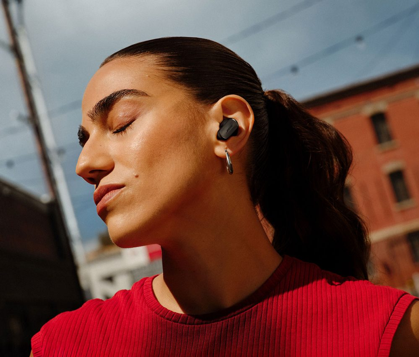 Mujer con ojos cerrados, auriculares, camiseta roja, pendiente plateado. Fondo: edificio y cielo.