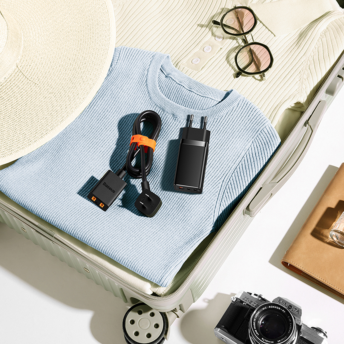 Otwarta walizka, spakowana ubraniami, ładowarką, kapeluszem, okularami przeciwsłonecznymi i aparatem. Białe tło. Gotowe do podróży.