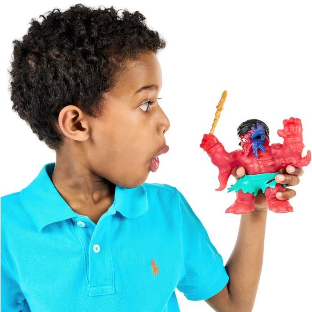 Ein Junge betrachtet ein rotes Monstermonsterspielzeug. Der Junge trägt ein blaues Hemd. Das Spielzeug hat einen Speer und grüne Shorts.