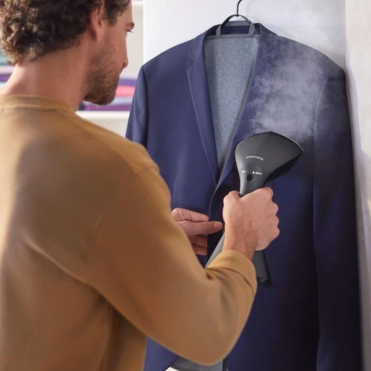 Mężczyzna paruje granatową marynarkę czarnym parownikiem. Para unosi się z marynarki. Mężczyzna ma na sobie brązową koszulę.