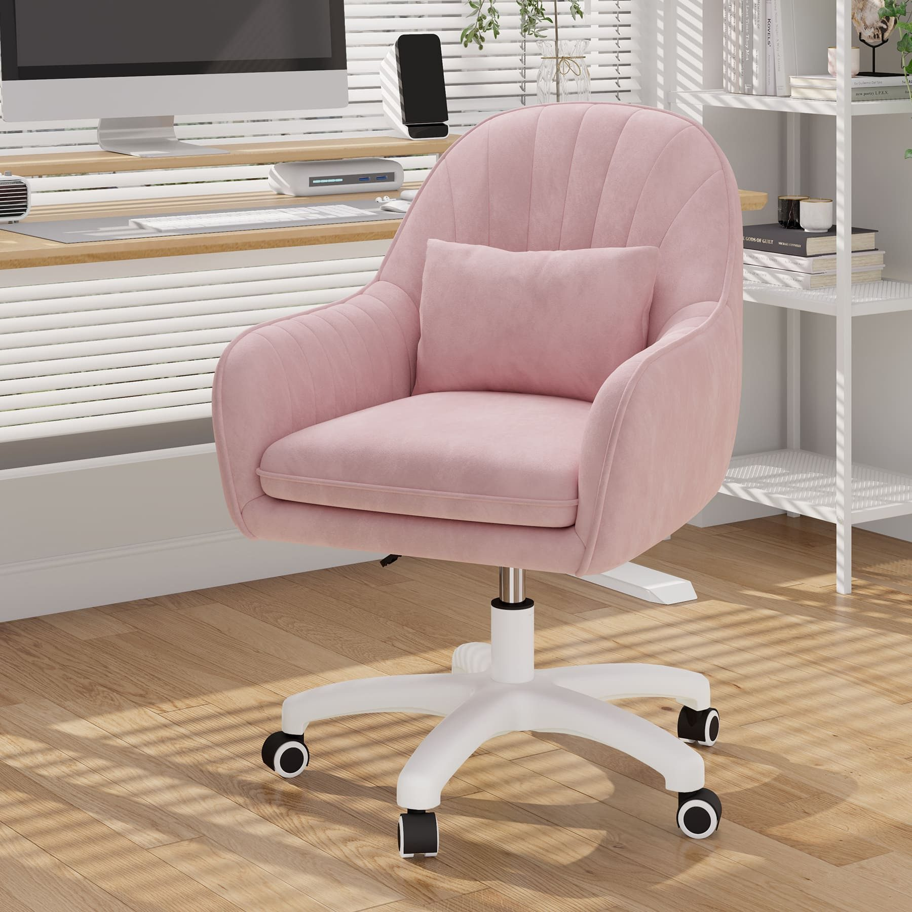 Una silla de oficina rosa con una almohada frente a un escritorio. Una computadora, estanterías con libros y persianas en el fondo.