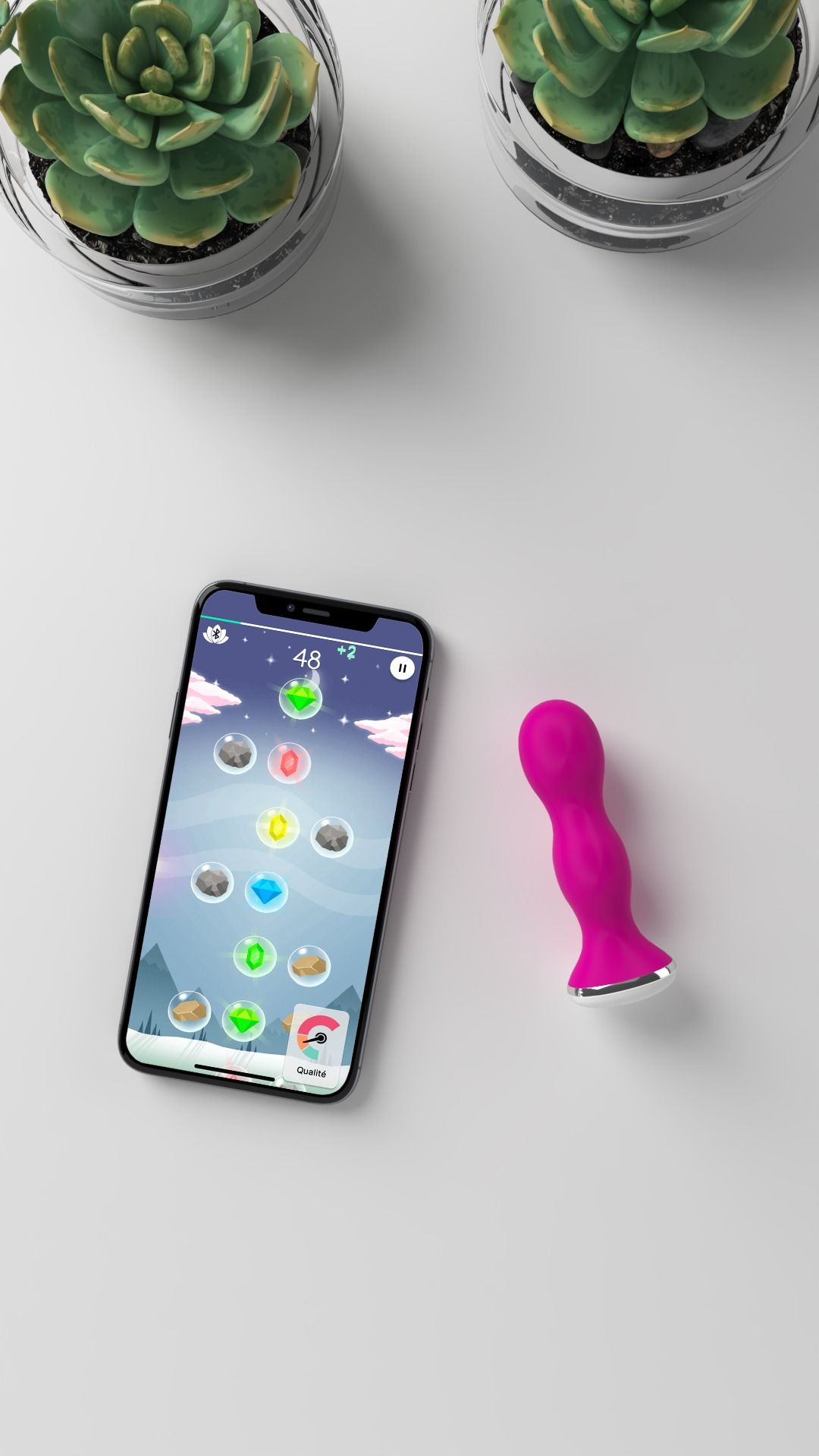 Ein Telefon mit einem Spiel, ein rosa Vibrator und zwei Sukkulenten in Glastöpfen auf einer weißen Oberfläche.