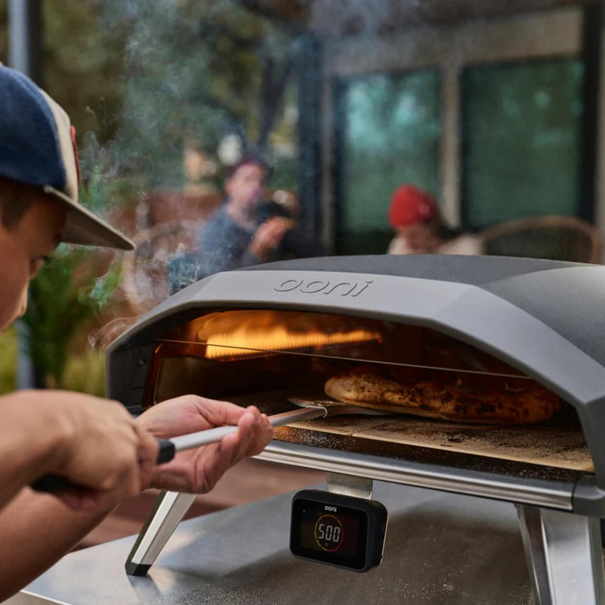 Eine Person holt eine Pizza aus einem Pizzaofen. Der Ofen steht auf einem Tisch, und im Hintergrund sind Personen.