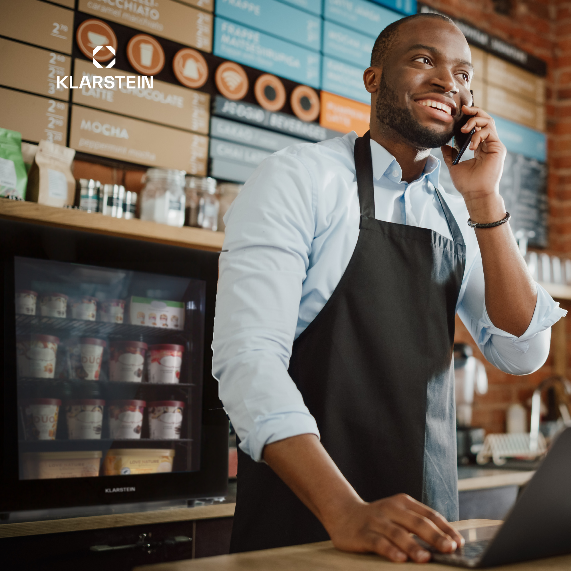 Ein lächelnder Barista in einem Café telefoniert und blickt auf einen Laptop. Hinter ihm sind Menütafeln und ein Kühlschrank mit Eis.