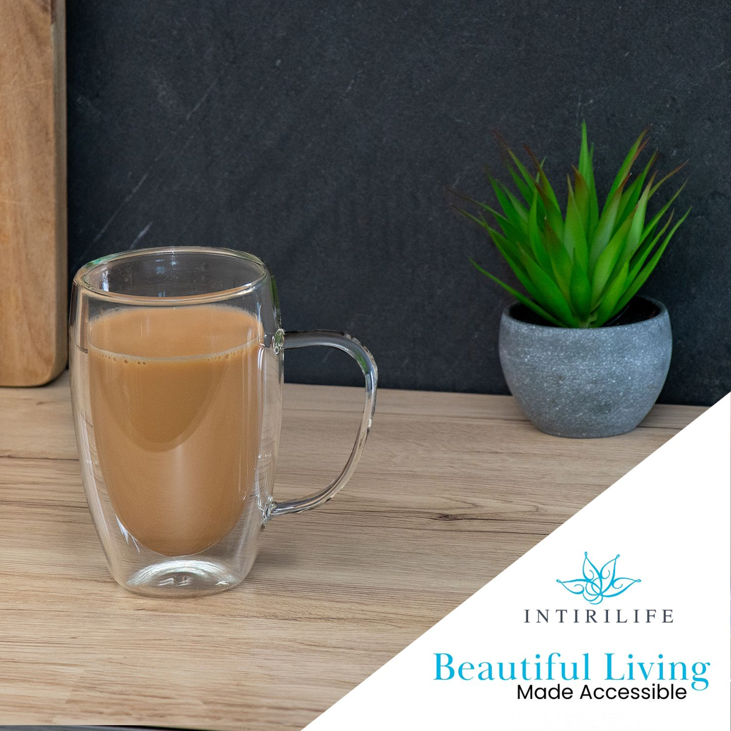 Eine klare Glastasse mit Kaffee steht auf Holz. Daneben steht eine kleine Pflanze in einem grauen Topf. Links ist ein Holzbrett.
