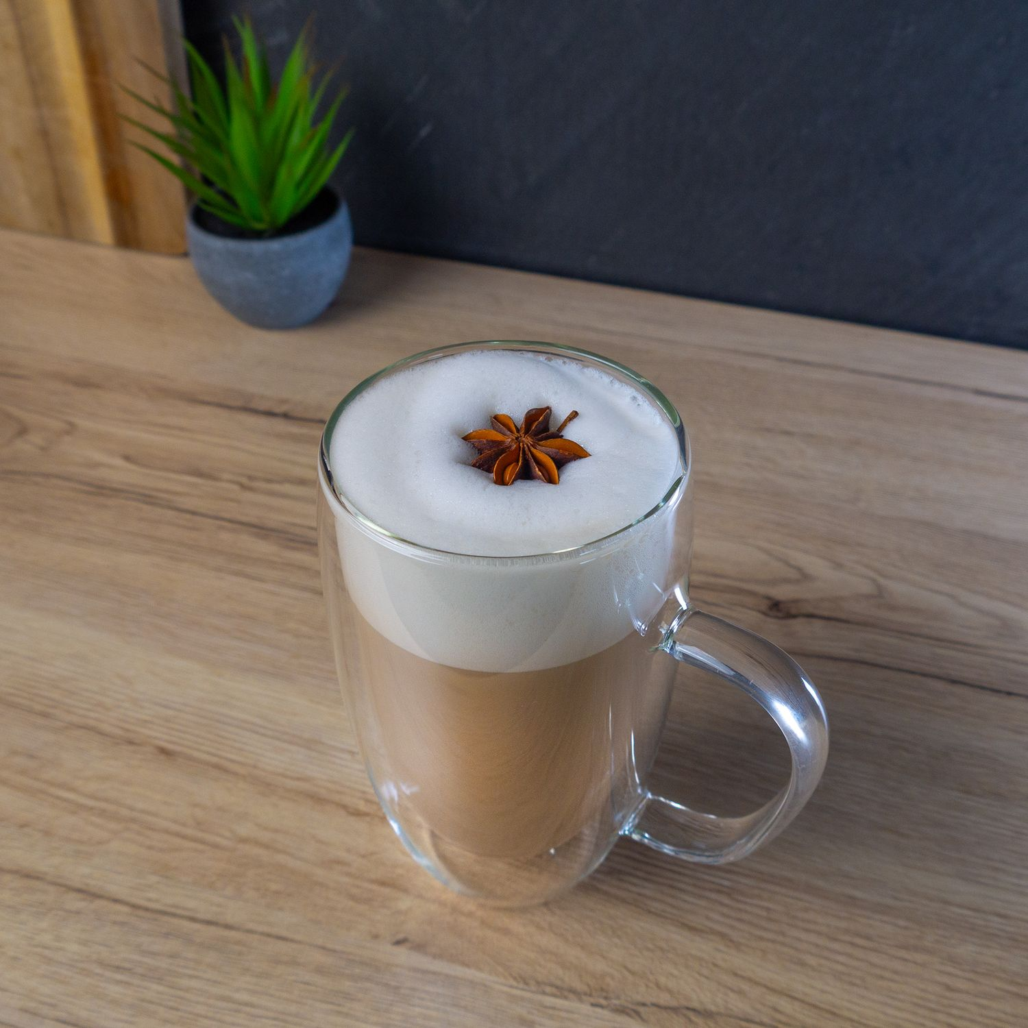 Ein klares Glas mit Latte, bedeckt mit Schaum und Sternanis. Es steht auf einer Holzoberfläche, im Hintergrund eine kleine Pflanze.