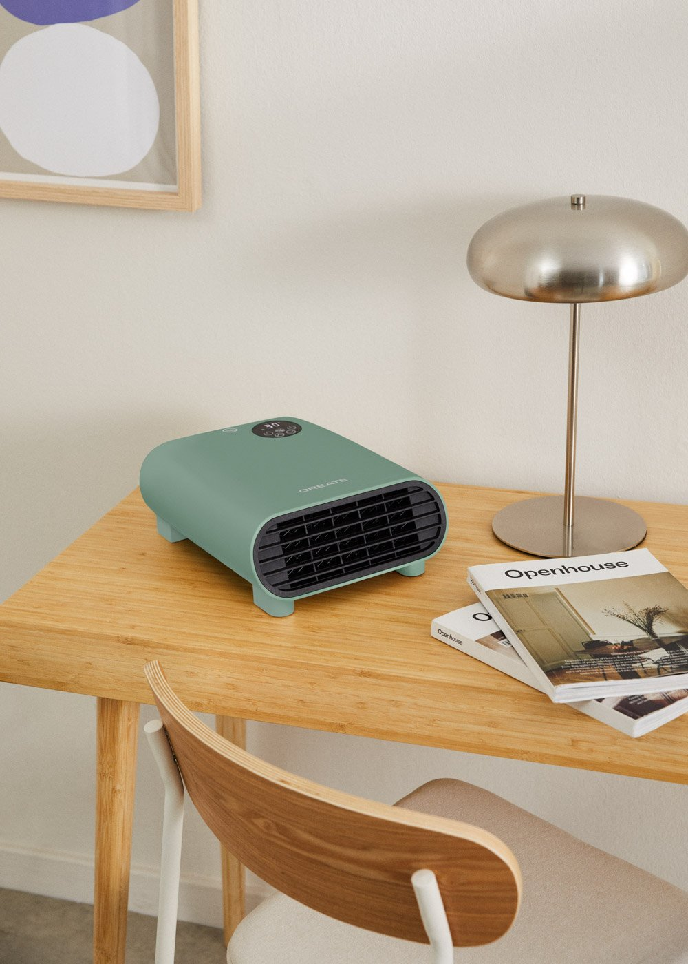 Un calentador verde menta está sobre un escritorio de madera con una lámpara, libros y una silla. Un cuadro cuelga en la pared.