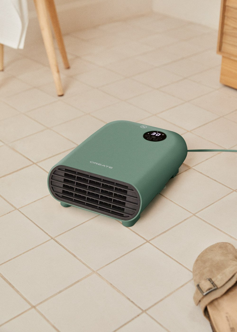 Un calefactor verde con pantalla está sobre baldosas. Una sandalia está cerca. Patas de madera y un armario al fondo.