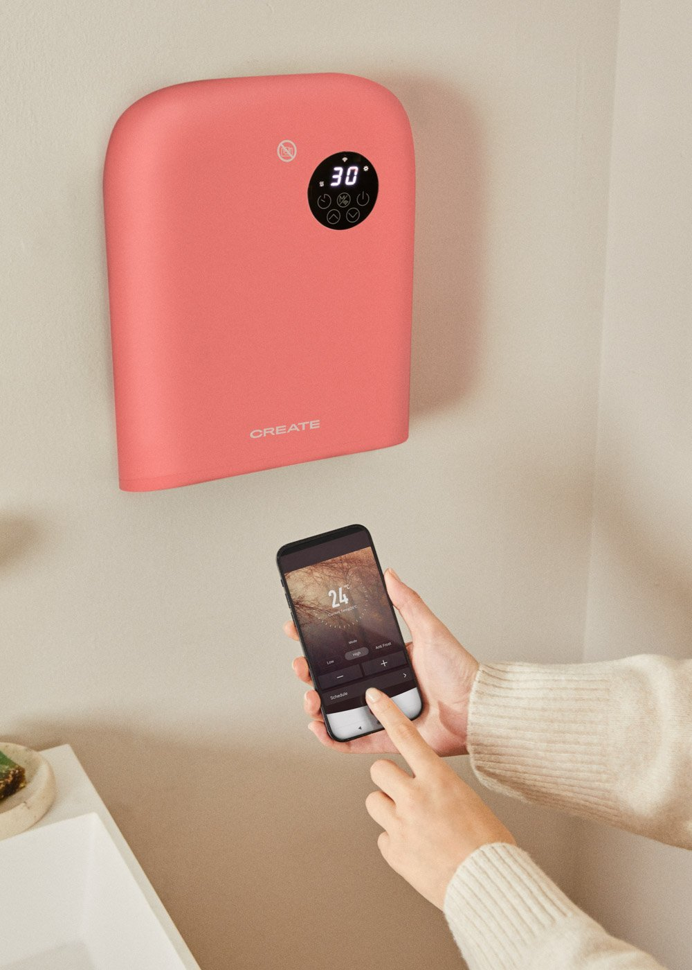 Una persona usa un teléfono para controlar un calentador de pared rosa. El calentador muestra 30 grados. El teléfono muestra 24 grados.